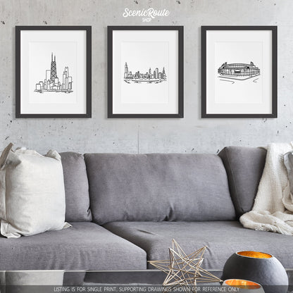 A group of three framed drawings on a wall hanging above a couch with pillows and a blanket. The line art drawings include the John Hancock Building, the Chicago Skyline, and Wrigley Field