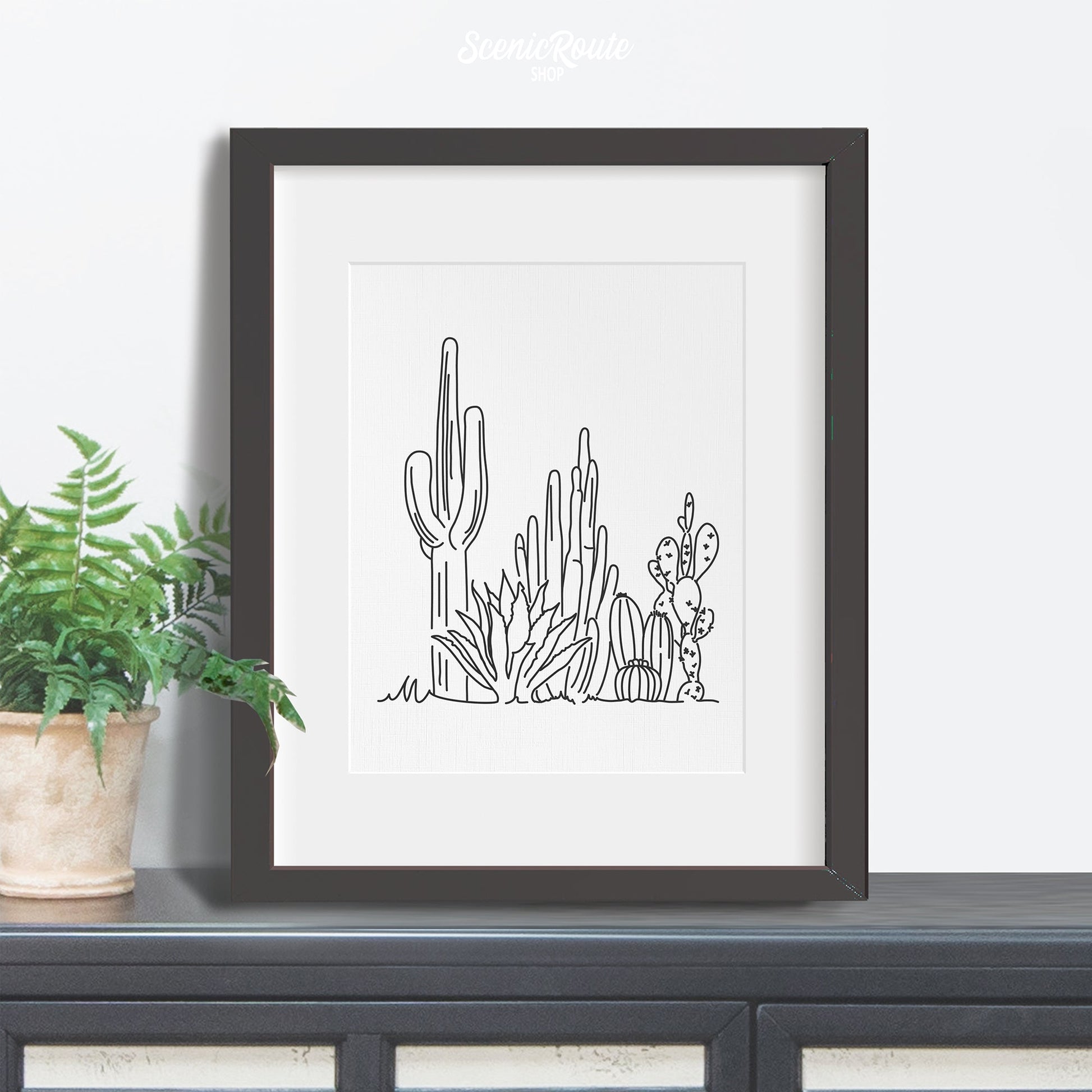 A framed line art drawing of a Cactus Garden on a credenza with plant