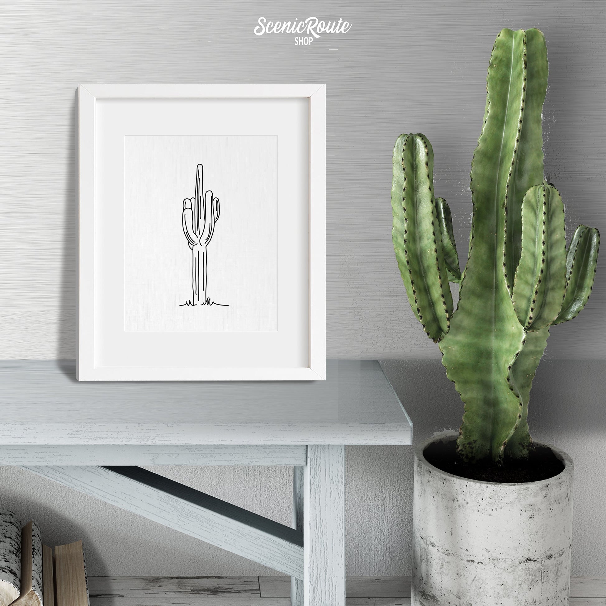 A framed line art drawing of a Saguaro Cactus sitting on a bench next to a potted cactus