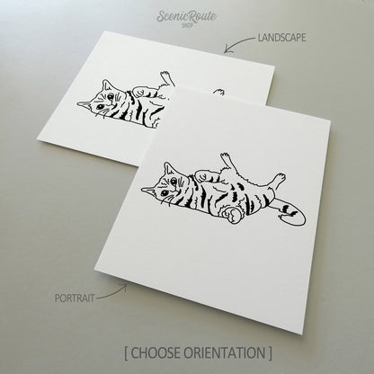 Two line art drawings of a Playful Cat on white linen paper with a gray background.  The pieces are shown in portrait and landscape orientation for the available art print options.