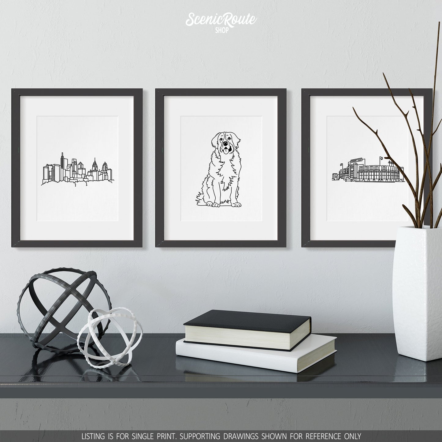 A group of three framed drawings on a white wall above a black table with books and a vase. The line art drawings include the Philadelphia Skyline, a Bernese Mountain Dog, and Lambeau Field