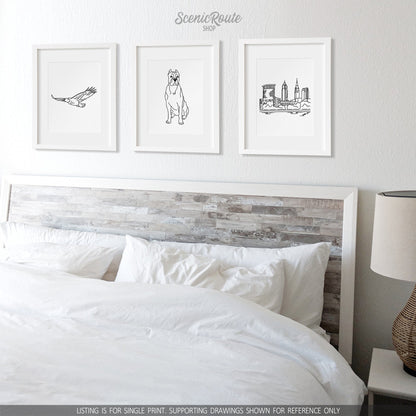 A group of three framed drawings on a white wall above a bed with white sheets and a nightstand with a lamp.  The line art drawings include an Eagle flying, a Cane Corso dog, and the Cleveland Skyline