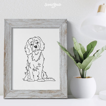 A framed line art drawing of a Cavalier King Charles Spaniel dog on a desk next to a lamp and a potted plant