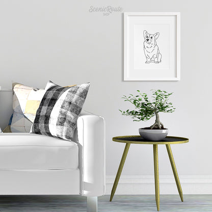 A framed line art drawing of a Corgi dog on a white wall hanging above a side table and plant next to a couch