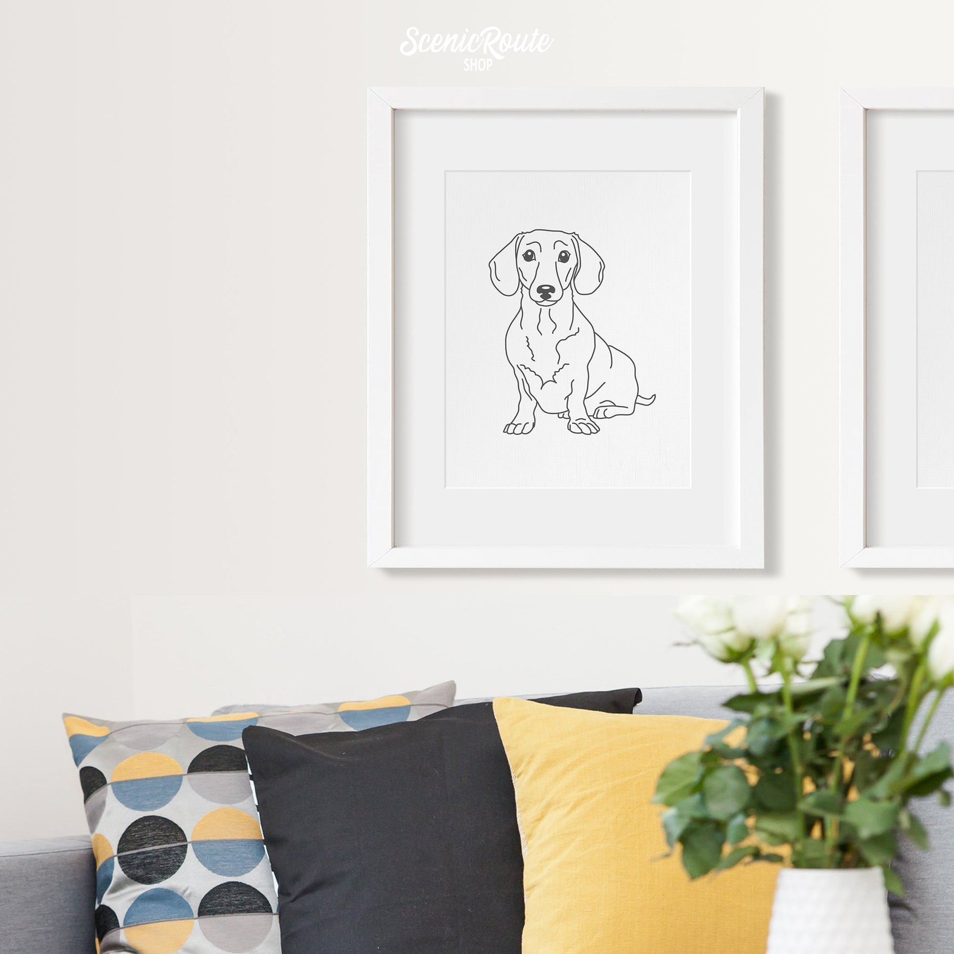 A framed line art drawing of a Dachshund dog on a white wall hanging above a couch with pillows