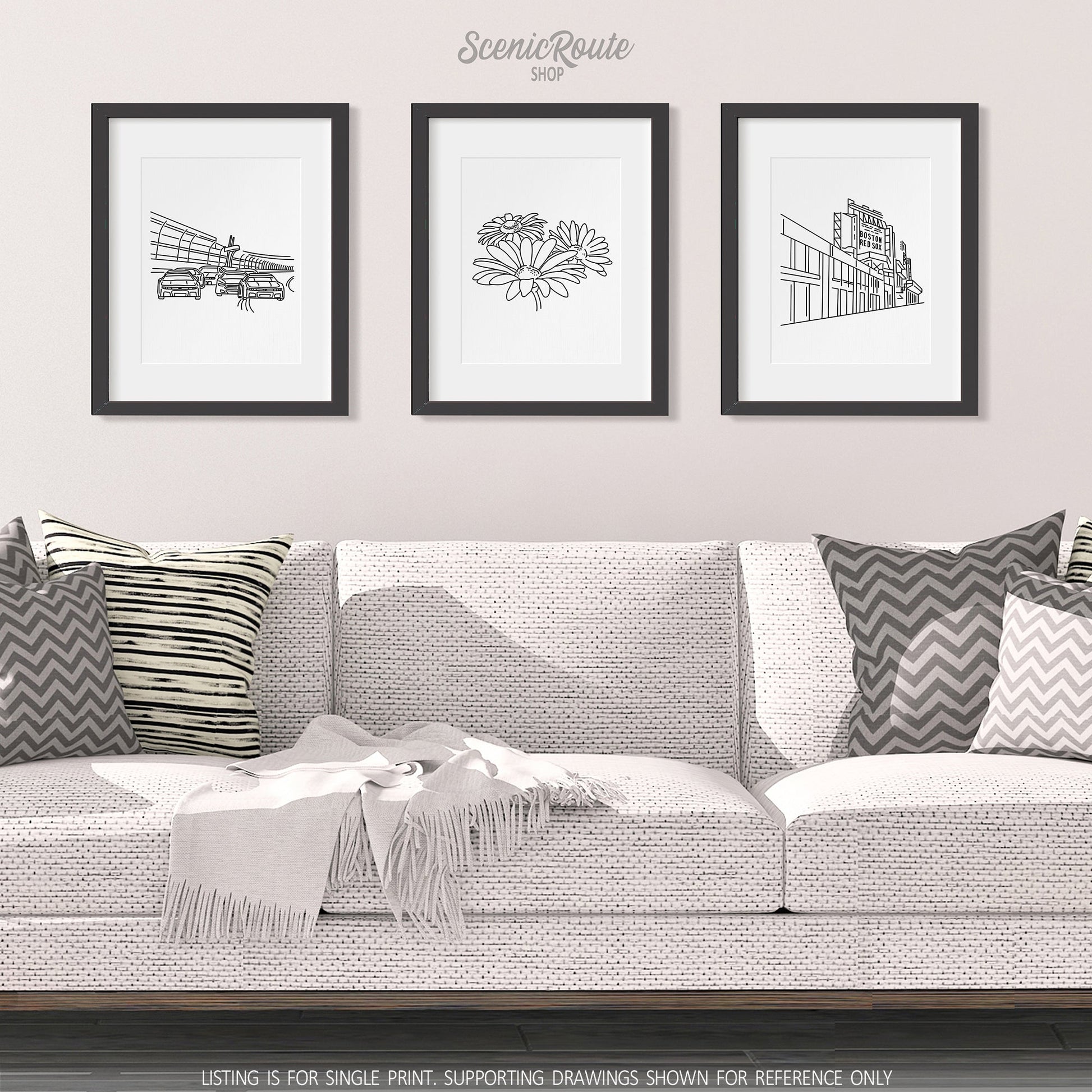 A group of three framed drawings on a white wall hanging above a couch with pillows and a blanket. The line art drawings include Nascar, a Daisy Flower, and Fenway Park