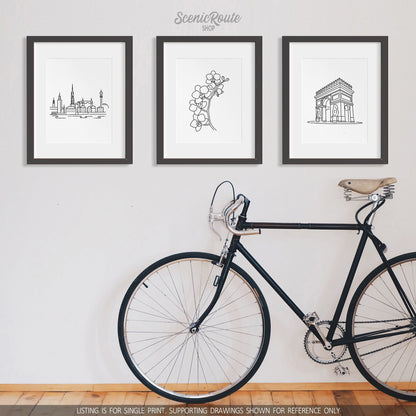 A group of three framed drawings on a white wall above a bicycle. The line art drawings include the Stockholm Skyline, an Orchid Flower, and the Paris Arc de Triomphe