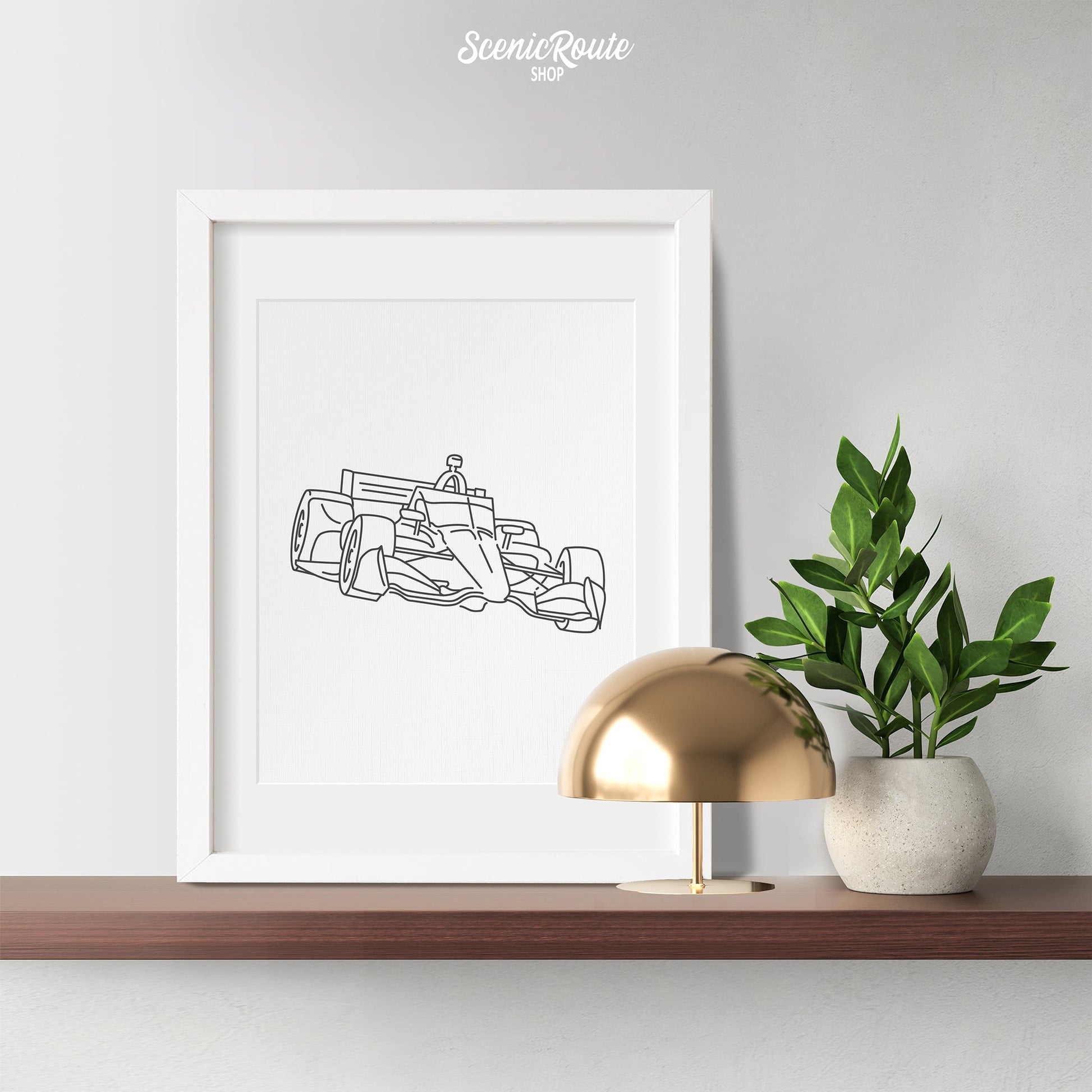 A framed line art drawing of an Indy Car on a wood shelf with a plant and lamp