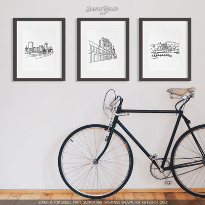 A group of three framed drawings on a white wall above a bicycle. The line art drawings include the Boston Skyline, Fenway Park, and Harvard Boathouse