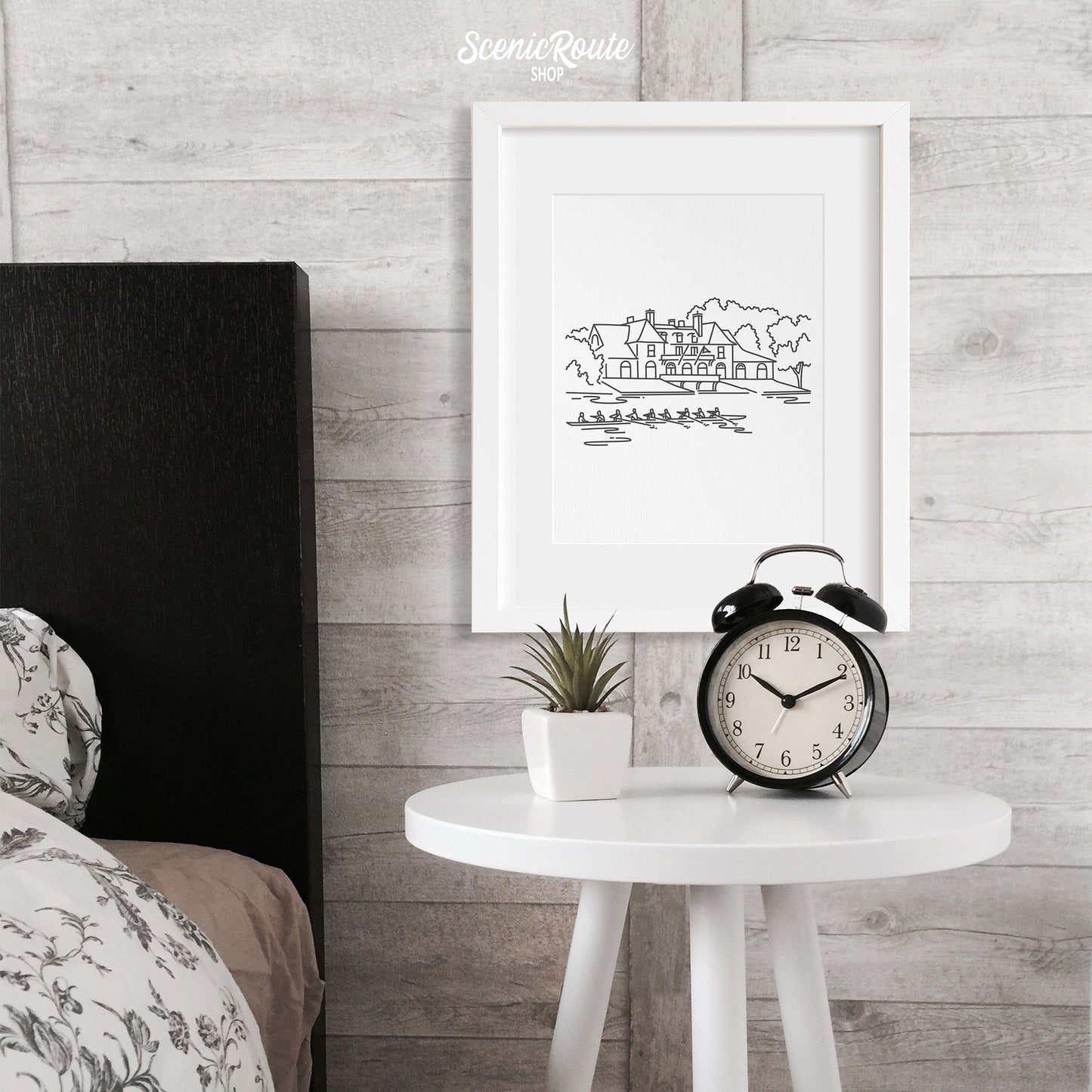 A framed line art drawing of the Harvard Boathouse above a nightstand next to a bed