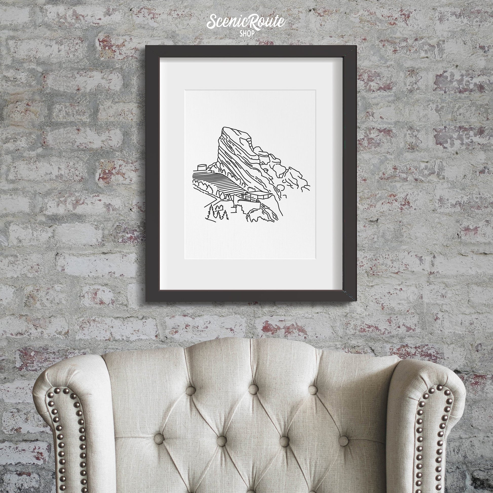 A framed line art drawing of the Red Rocks Amphitheatre on a brick wall above a chair