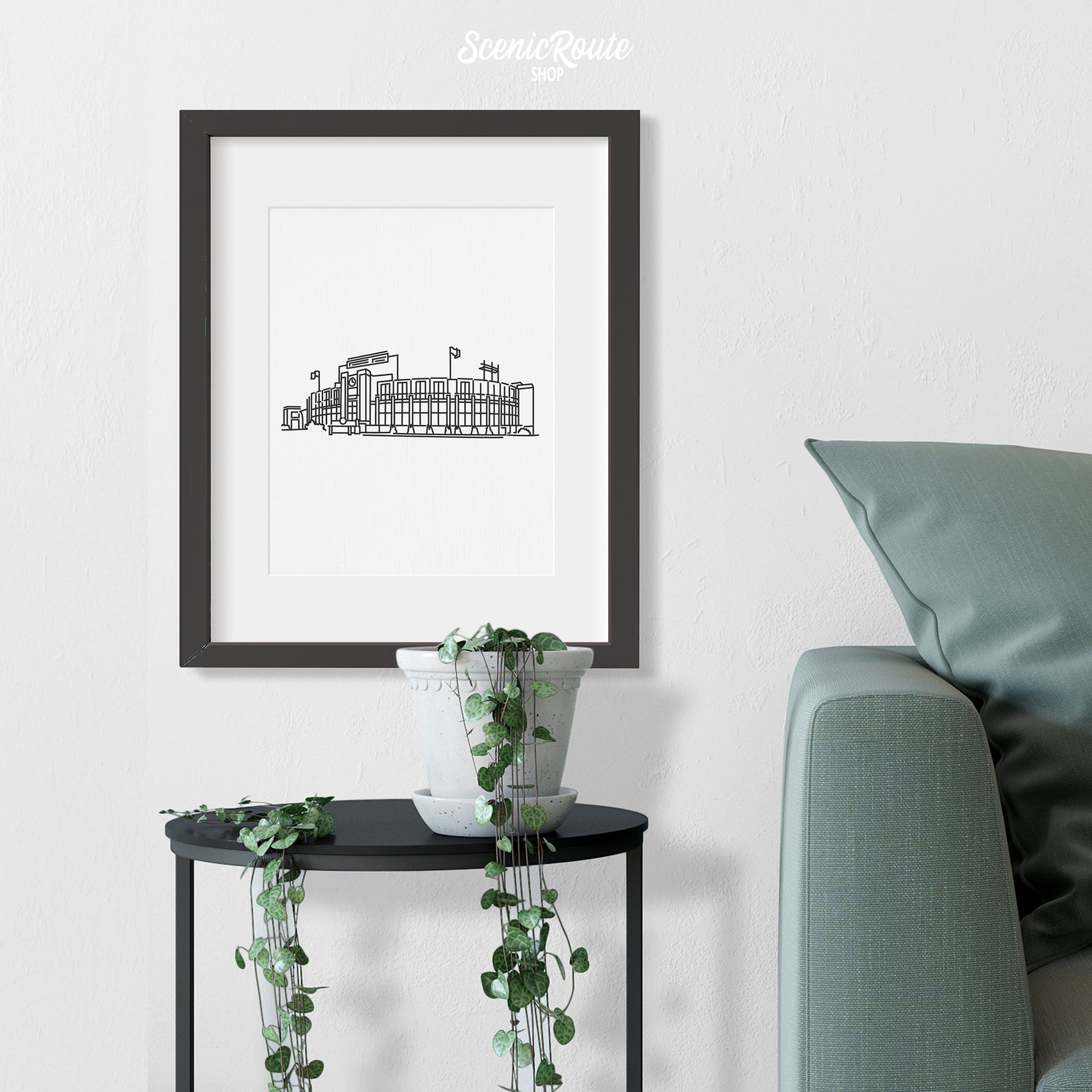A framed line art drawing of Lambeau Field above a side table with a plant