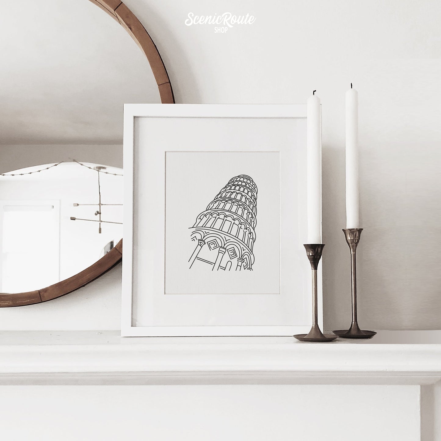 A framed line art drawing of the Leaning Tower of Pisa on a fireplace mantle with candles and a mirror