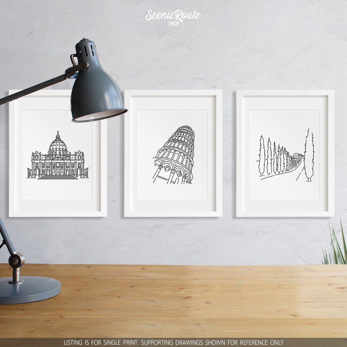 A group of three framed drawings on a wall above a desk with a lamp. The line art drawings include Saint Peters Basilica, the Leaning Tower of Pisa, and a Tuscany Drive