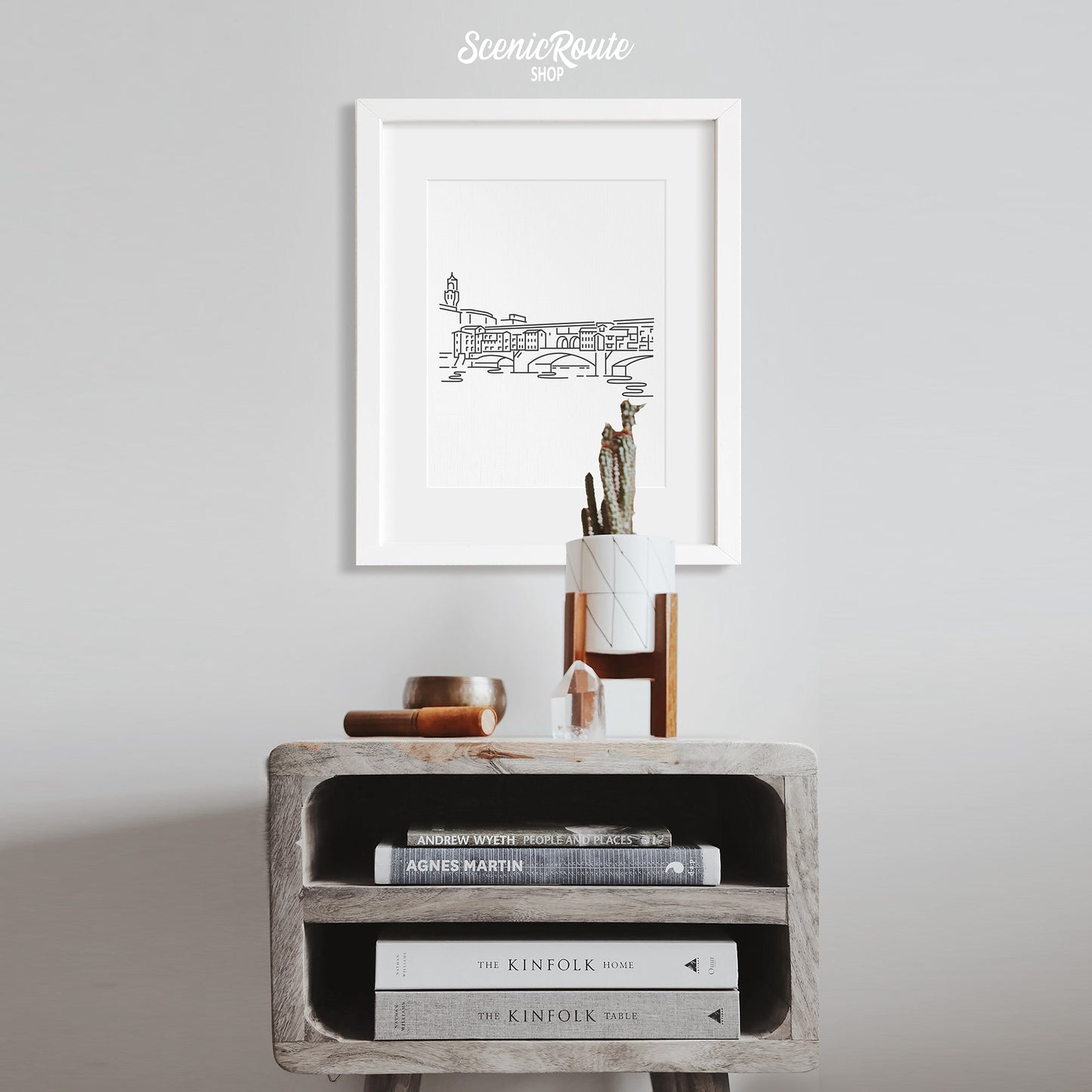 A framed line art drawing of the Ponte Vecchio hanging above a side table with a potted cactus