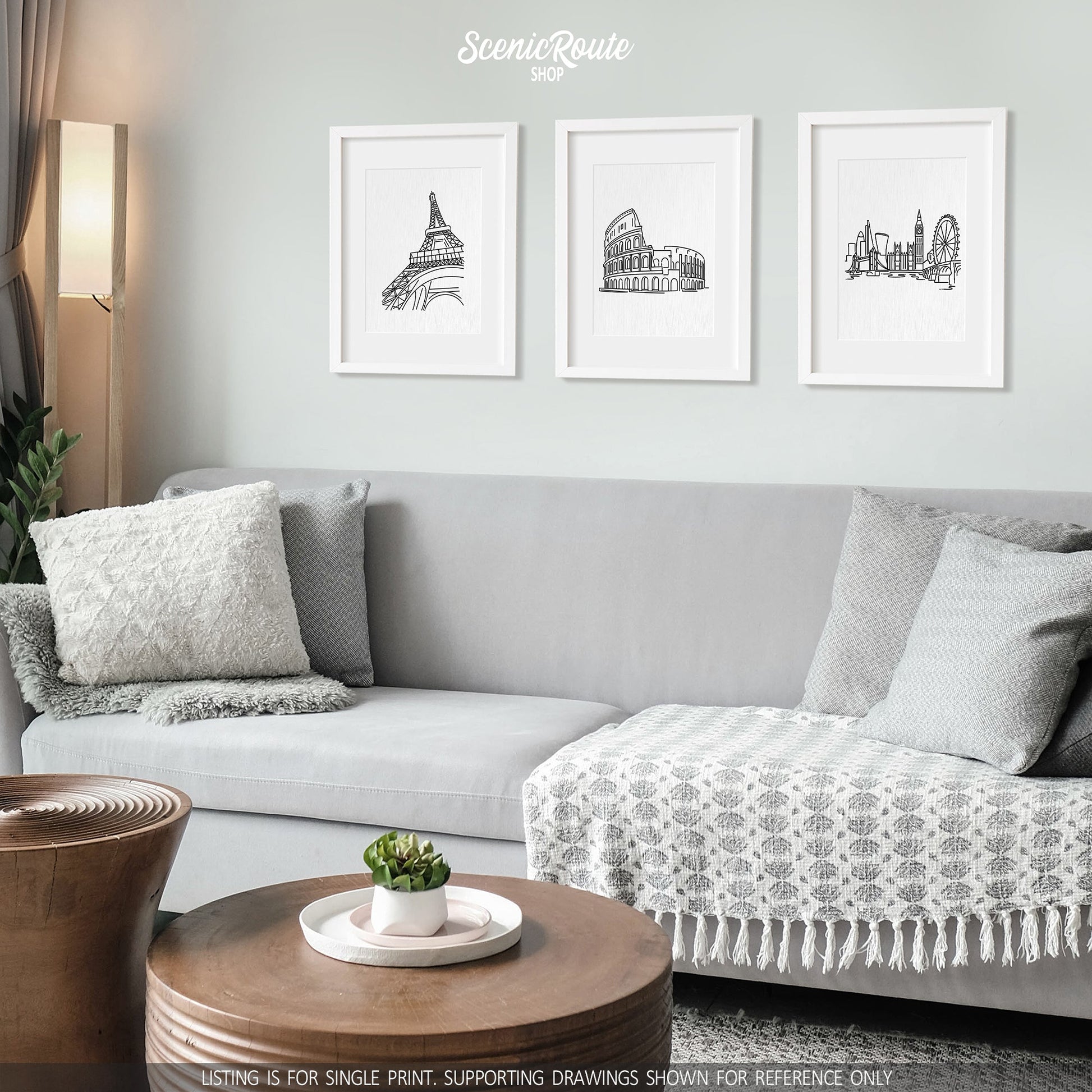 A group of three framed drawings on a white wall hanging above a couch with pillows and a blanket. The line art drawings include the Eiffel Tower, Colosseum, and the London Skyline