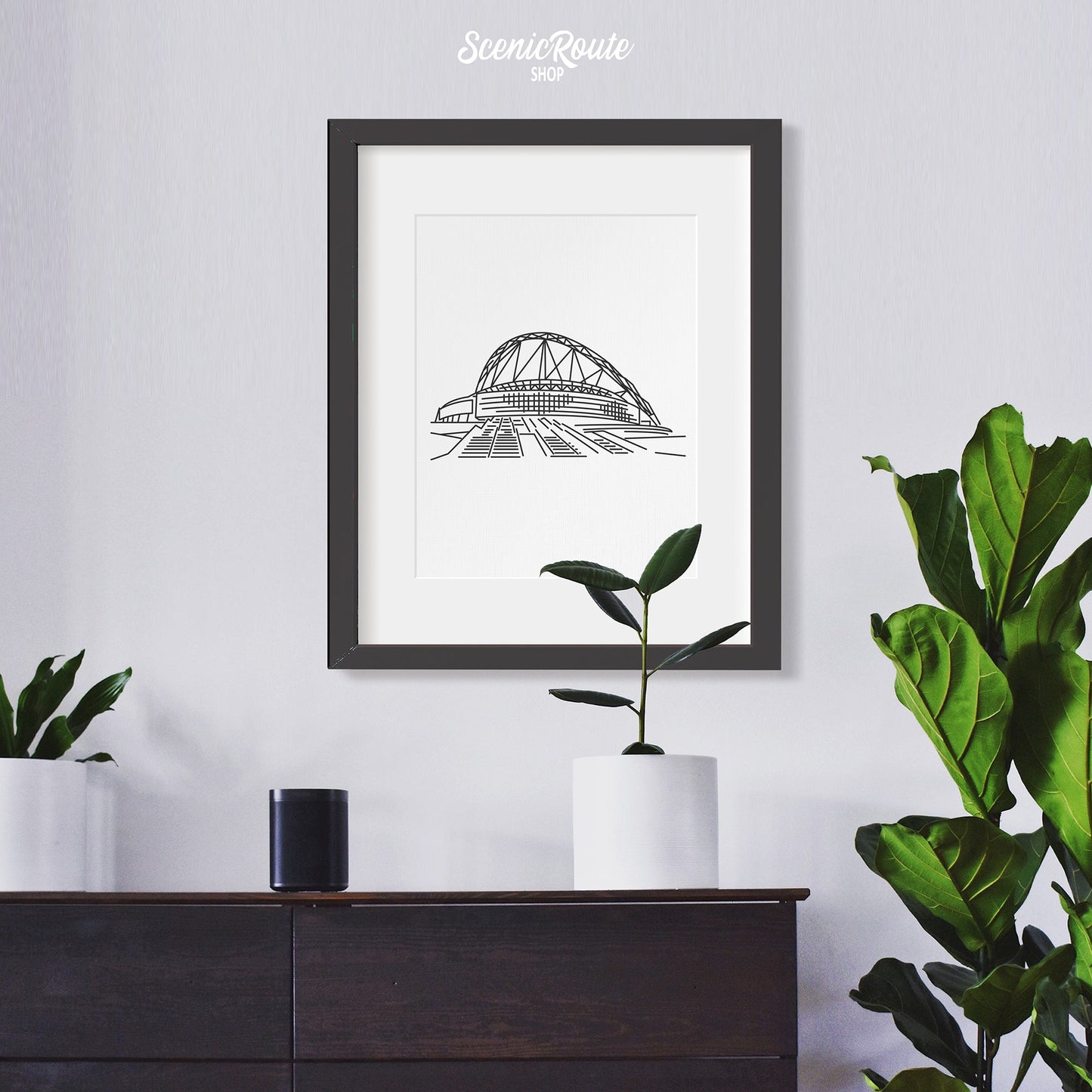 A framed line art drawing of Wembley Stadium above a dresser with plants