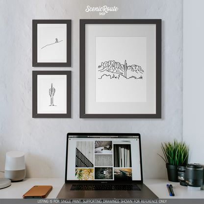 A group of three framed drawings on a wall above a desk and a laptop. The line art drawings include a person Hiking, a Saguaro Cactus, and the Superstition Mountains