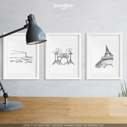 A group of three framed drawings on a wall above a desk with a lamp. The line art drawings include a Lighthouse, Drums, and the Eiffel Tower