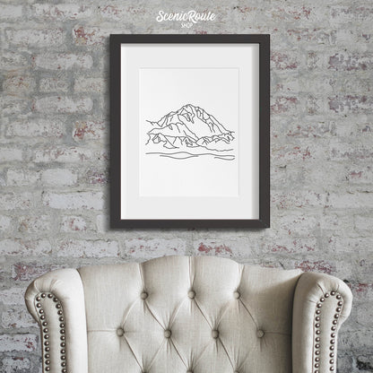 A framed line art drawing of Denali National Park on a brick wall above a chair