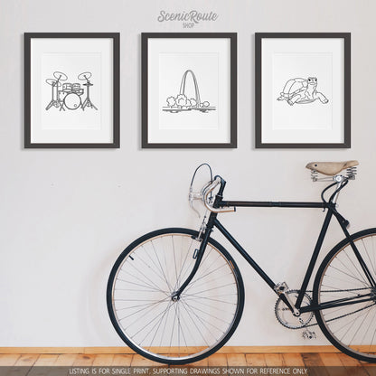 A group of three framed drawings on a white wall with a bicycle. The line art drawings include Drums, Gateway Arch National Park, and a Tortoise