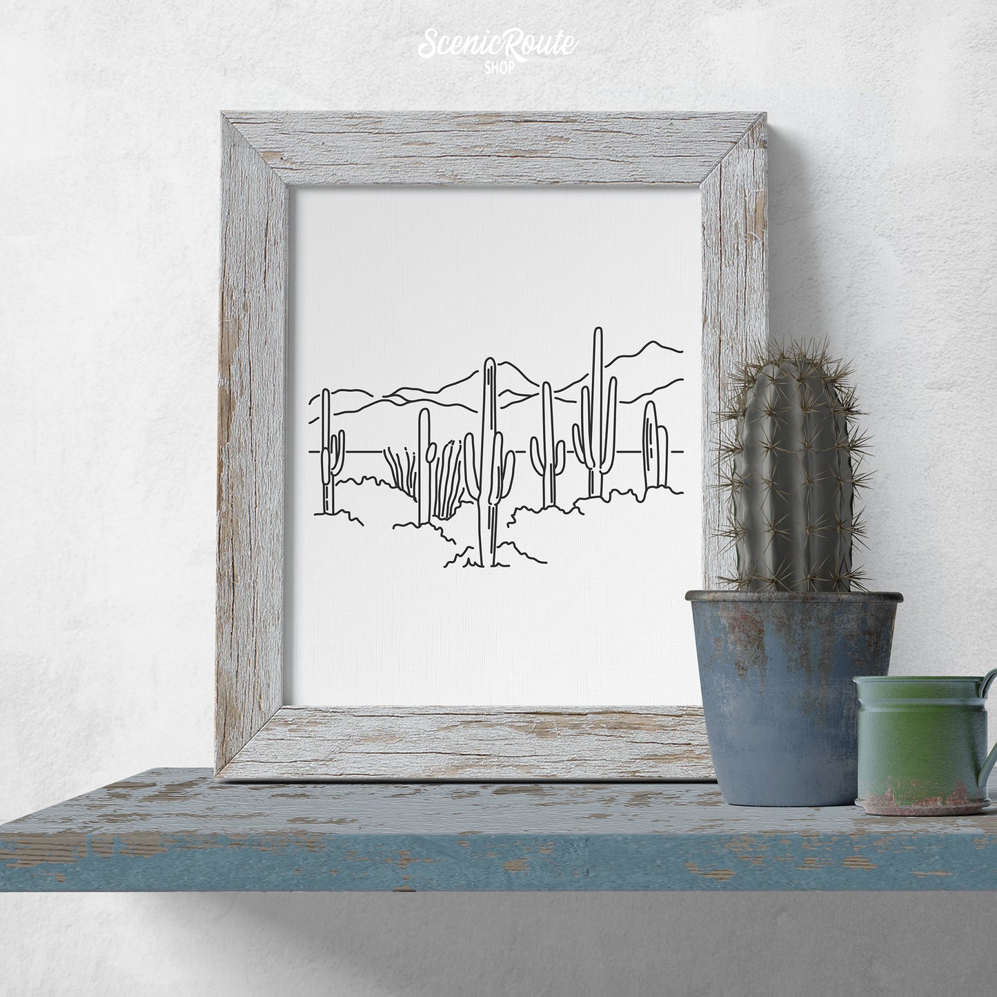 A framed line art drawing of Saguaro National Park on a blue shelf with a potted cactus