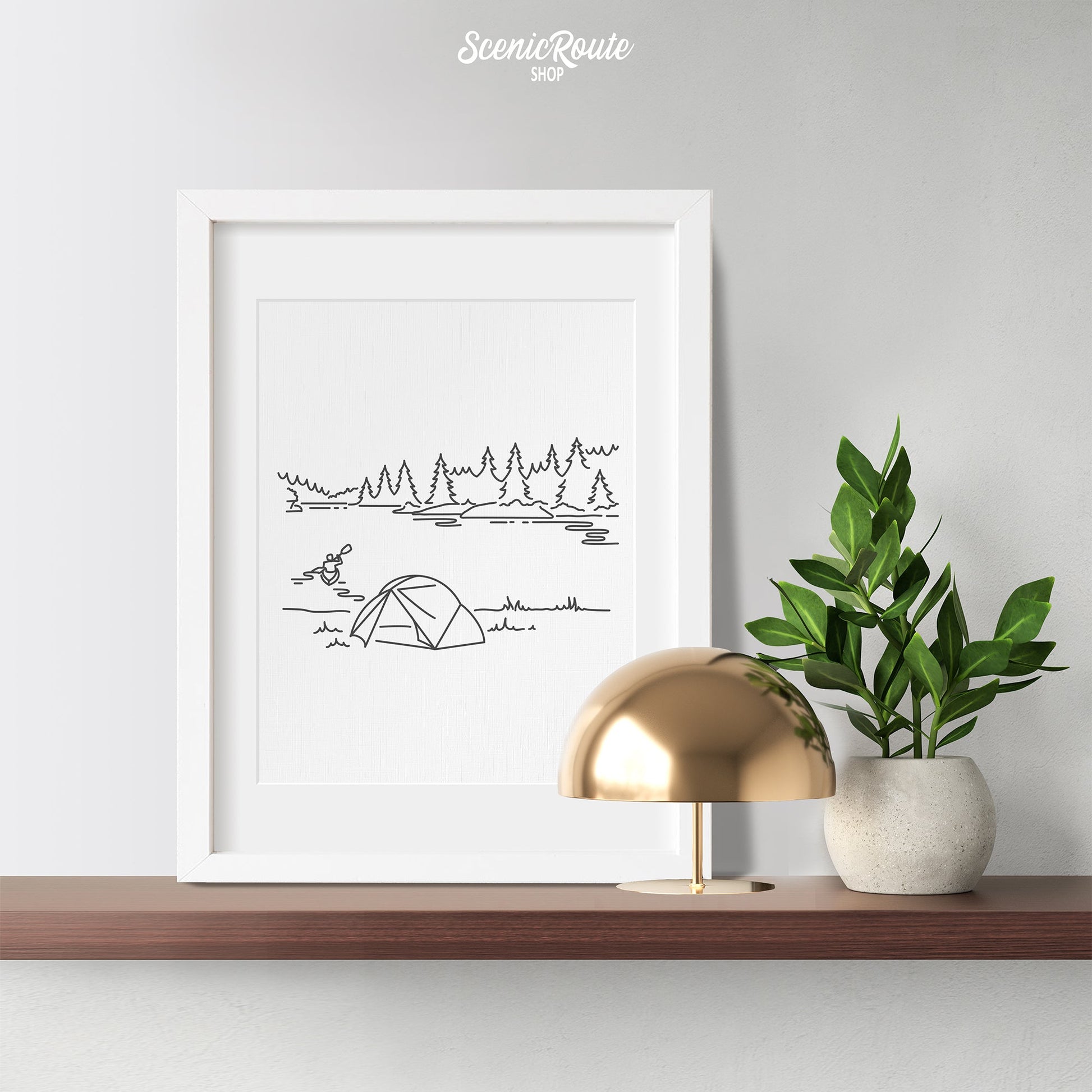 A framed line art drawing of Voyageurs National Park on a wood shelf with a lamp and plant