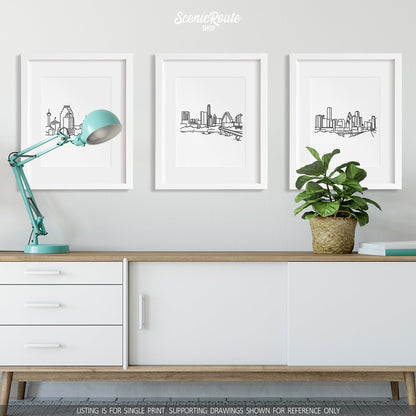 A group of three framed drawings on a wall hanging above a credenza with a lamp and a potted plant. The line art drawings include the San Antonio Skyline, Austin Skyline, and Houston Skyline