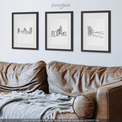 A group of three framed drawings on a wall above a leather couch with a blanket. The line art drawings include the Raleigh Skyline, Charlotte Skyline, and Shenandoah National Park