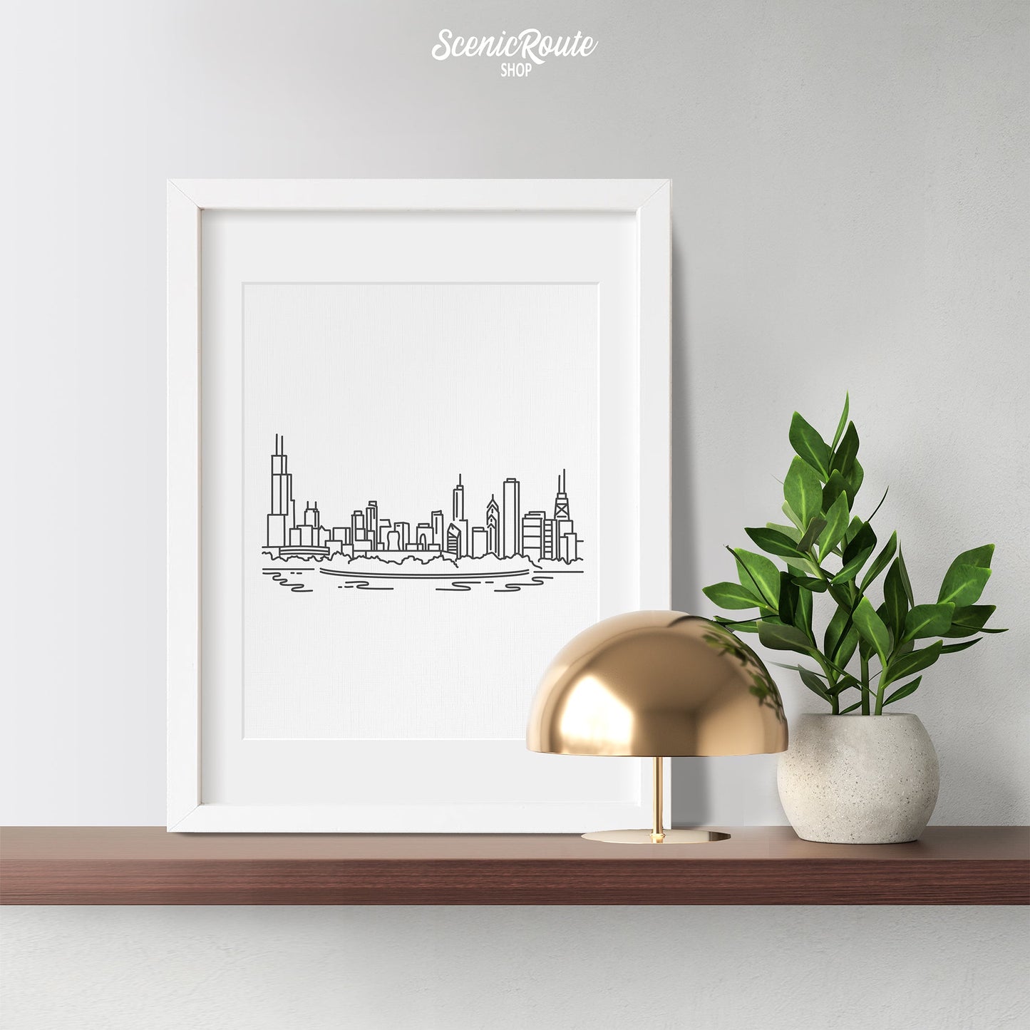 A framed line art drawing of the Chicago Skyline on a wood shelf with a plant and lamp