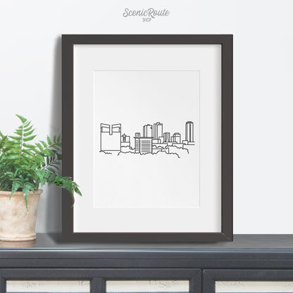 A framed line art drawing of the Fort Worth Skyline sitting on a credenza with a potted plant