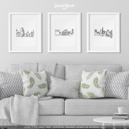 A group of three framed drawings on a white wall hanging above a couch with pillows and a blanket. The line art drawings include the San Antonio Skyline, Fort Worth Skyline, and Austin Skyline