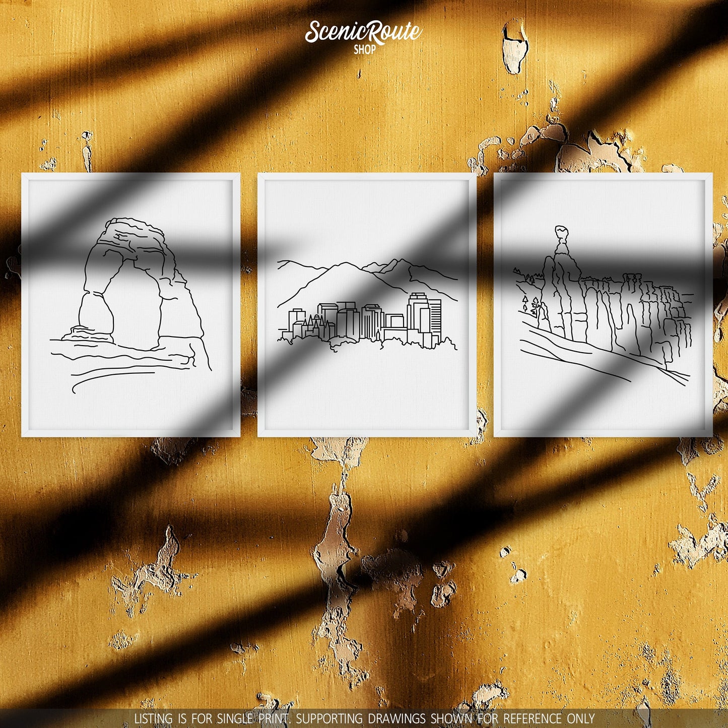A group of three framed drawings on a rough yellow wall with sun shadows. The line art drawings include Arches National Park, Salt Lake City Skyline, and Bryce Canyon National Park