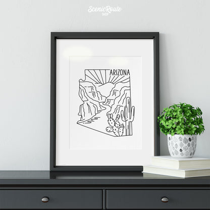 A framed line art drawing of the Arizona State Outline National Park on a dresser with books and a plant