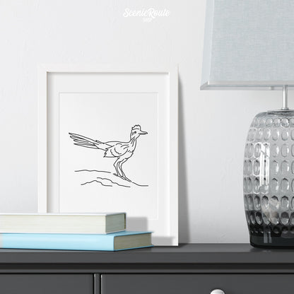 A framed line art drawing of a Roadrunner on a gray dresser with a lamp and books