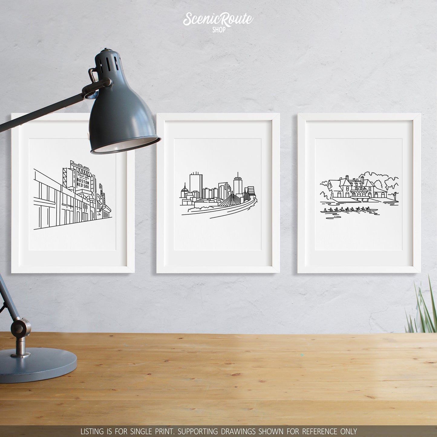 A group of three framed drawings on a wall above a desk with a lamp. The line art drawings include Fenway Park, the Boston Skyline, and the Harvard Boathouse