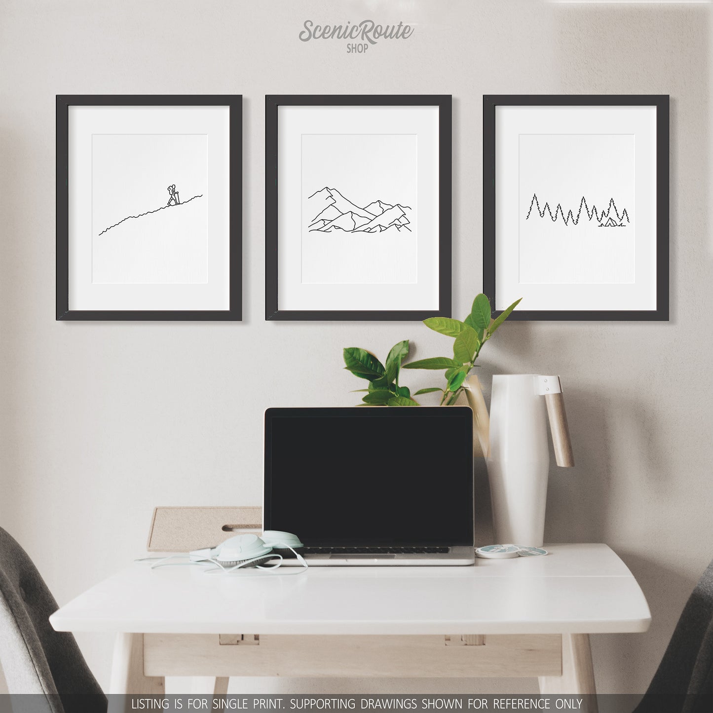 A group of three framed drawings on a wall above a desk with a laptop. The line art drawings include a person Hiking, a Mountain Range, and Camping