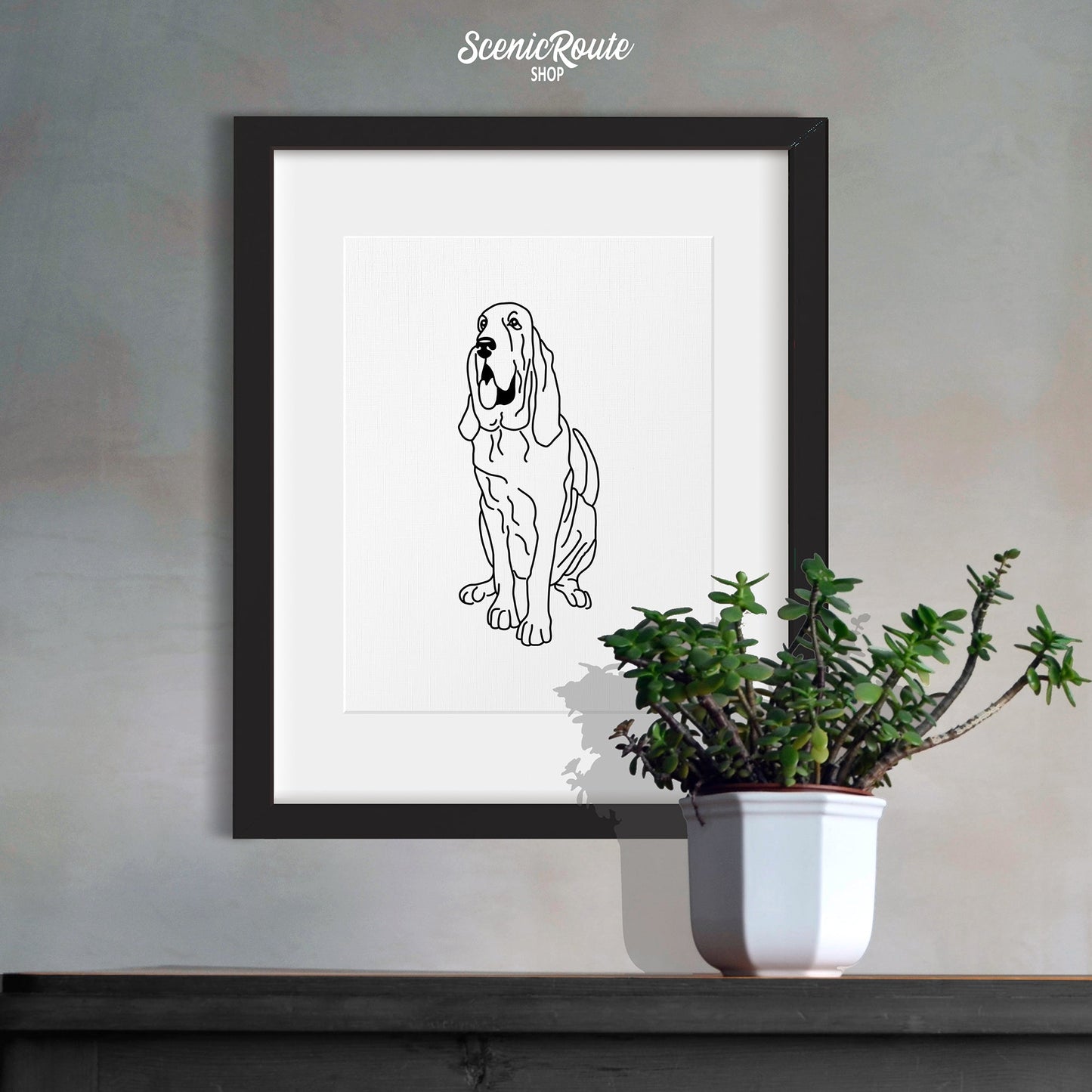 A framed line art drawing of a Bloodhound dog hanging above a gray cabinet with a plant on it