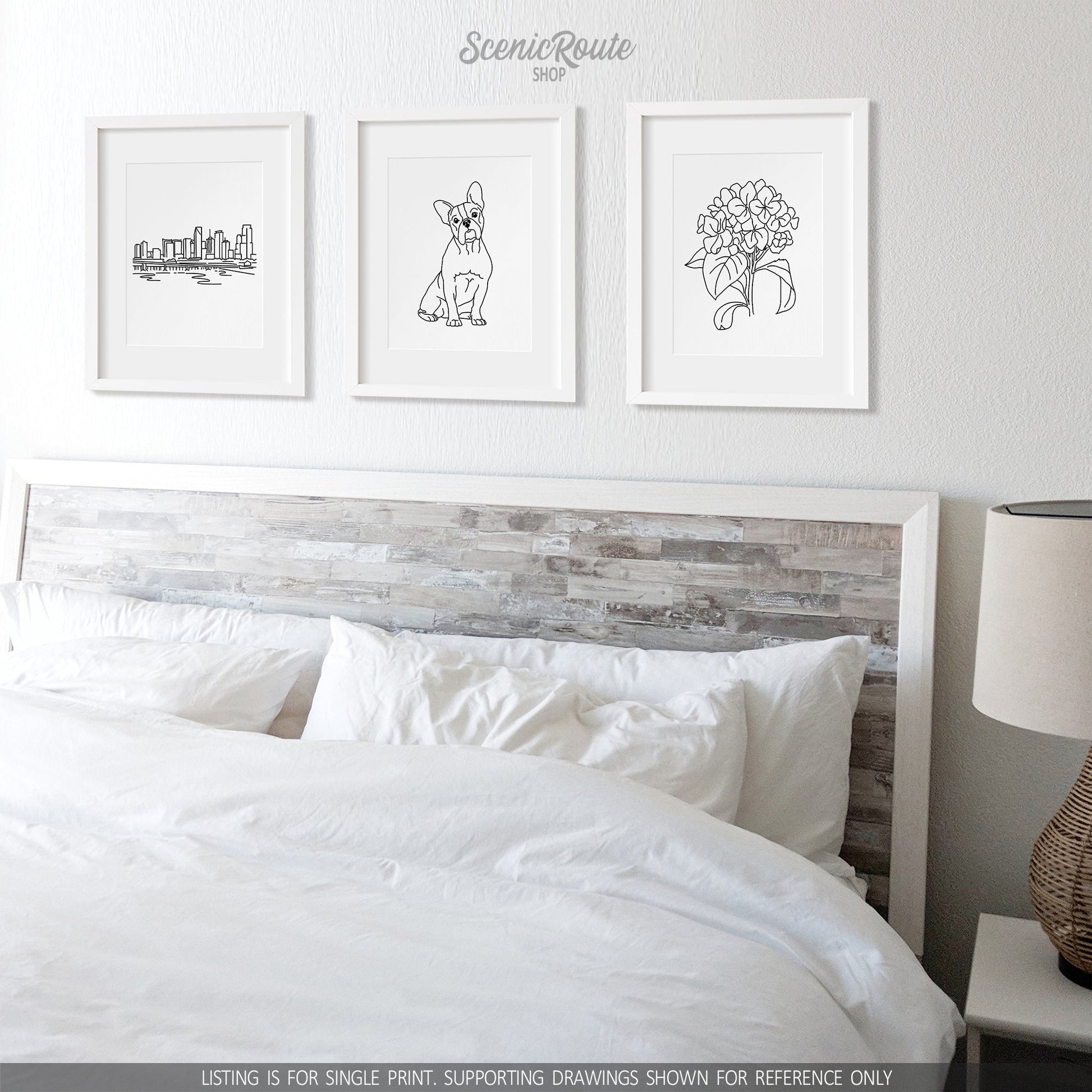 A group of three framed drawings on a white wall above a bed with white sheets and nightstand with a lamp. The line art drawings include the Miami Skyline, a Boston Terrier dog, and a Hydrangea Flower