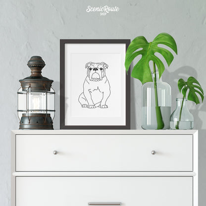 A framed line art drawing of a Bulldog on a white dresser next to a plant and lantern