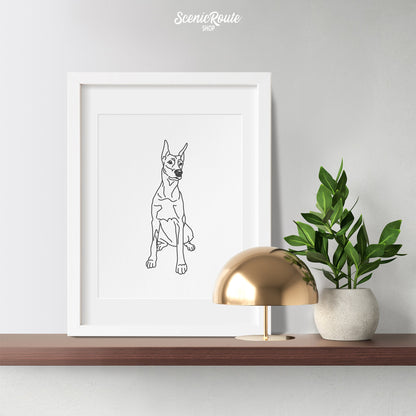 A framed line art drawing of a Doberman dog on a wood shelf with a plant and lamp