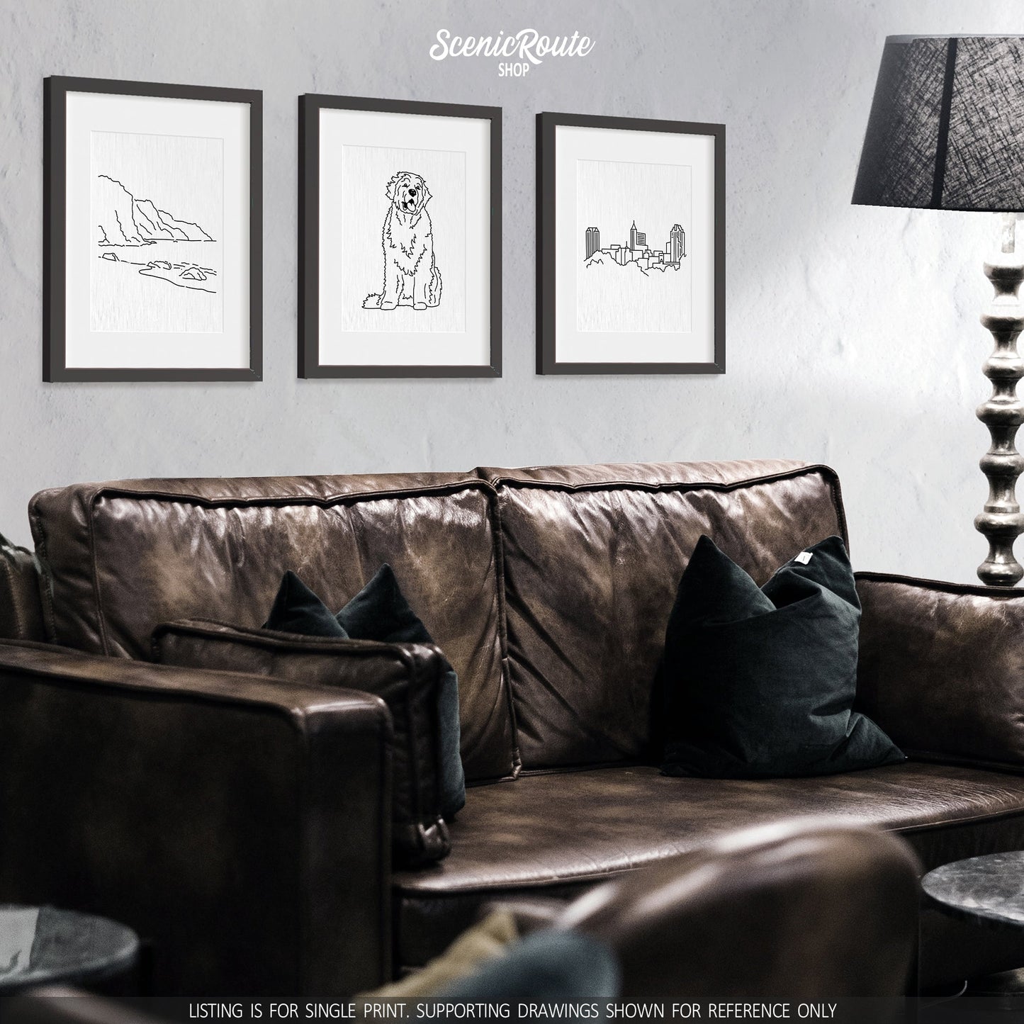 A group of three framed drawings on a white wall above a dark leather couch. The line art drawings include the NaPali Coast, a Newfoundland dog, and the Raleigh Skyline