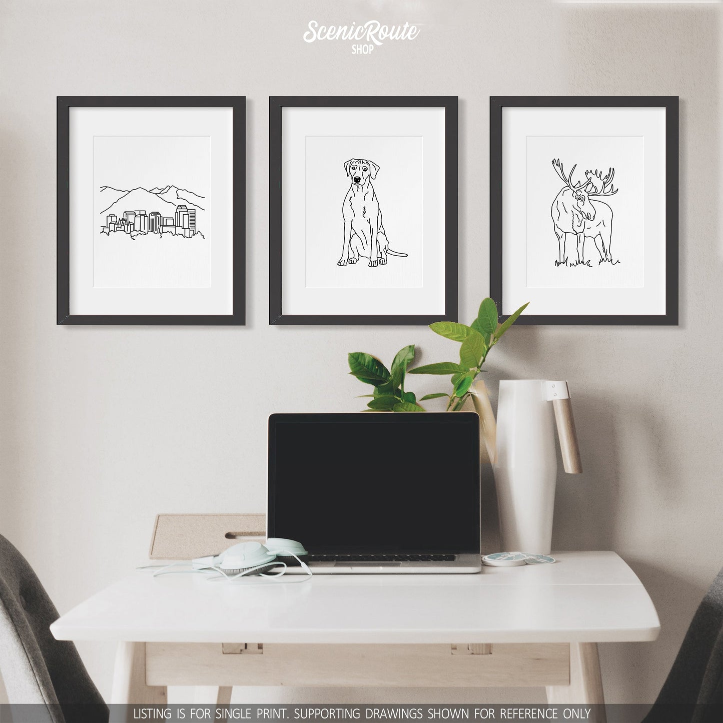 A group of three framed drawings on a white wall above a table with a laptop. The line art drawings include the Salt Lake City Skyline, a Rhodesian Ridgeback dog, and a Moose
