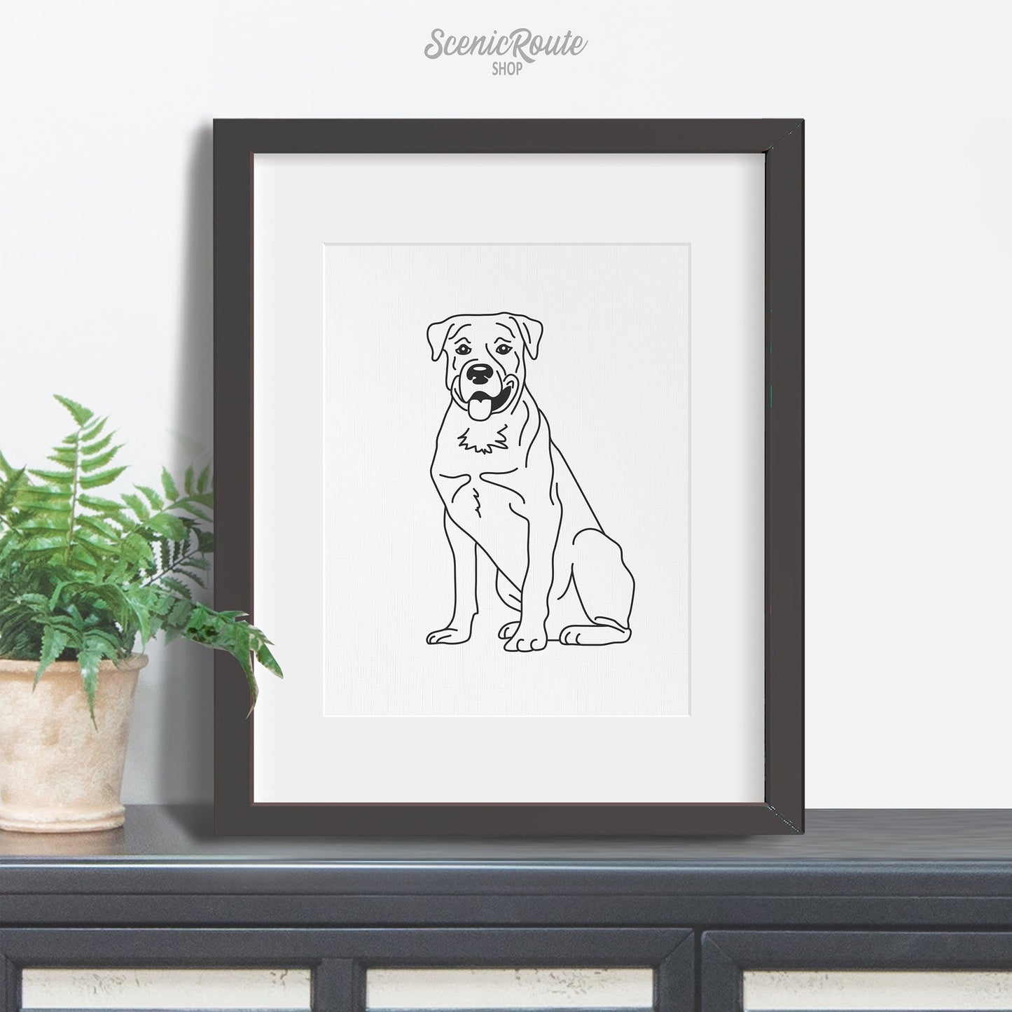 A framed line art drawing of a Rottweiler dog on a credenza with a plant
