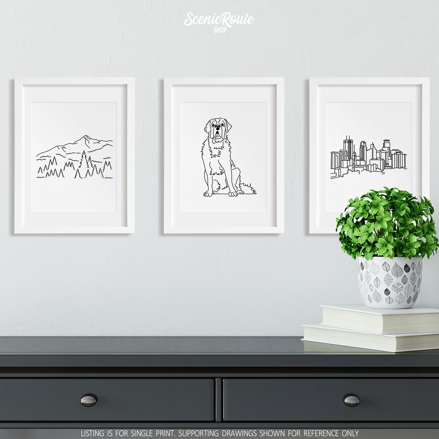 A group of three framed drawings on a white wall above a gray dresser with books and a plant. The line art drawings include Big Sky Montana, a Saint Bernard Dog, and the Minneapolis Skyline