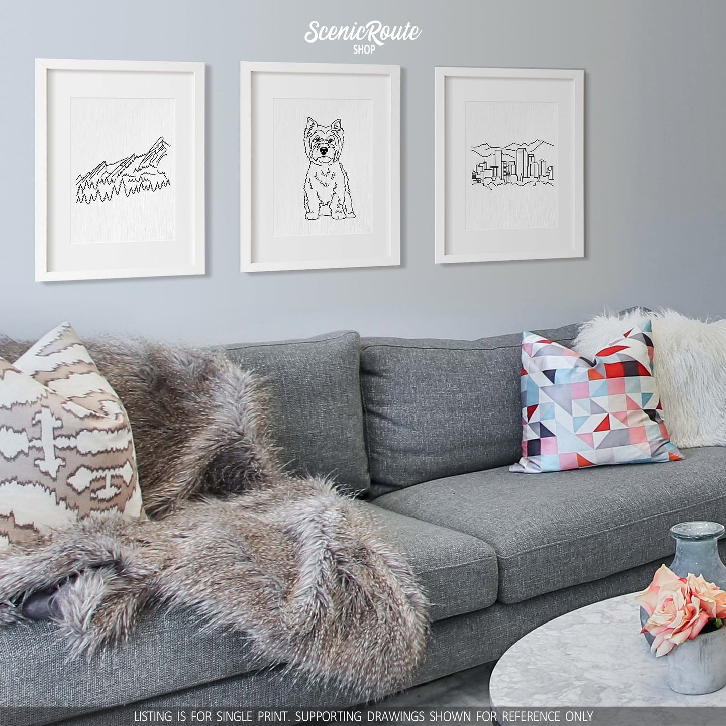 A group of three framed drawings on a white wall hanging above a couch with pillows and a blanket. The line art drawings include the Flatiron Mountains, a West Highland Terrier dog, and the Denver Skyline