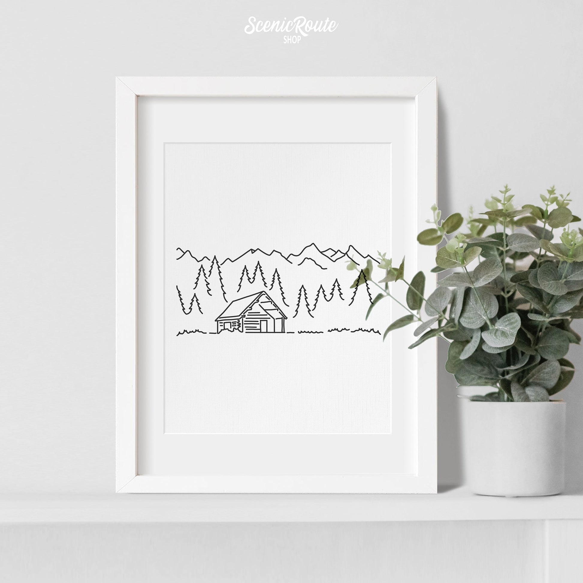 A framed line art drawing of a Log Cabin with a plant