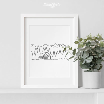 A framed line art drawing of a Log Cabin with a plant