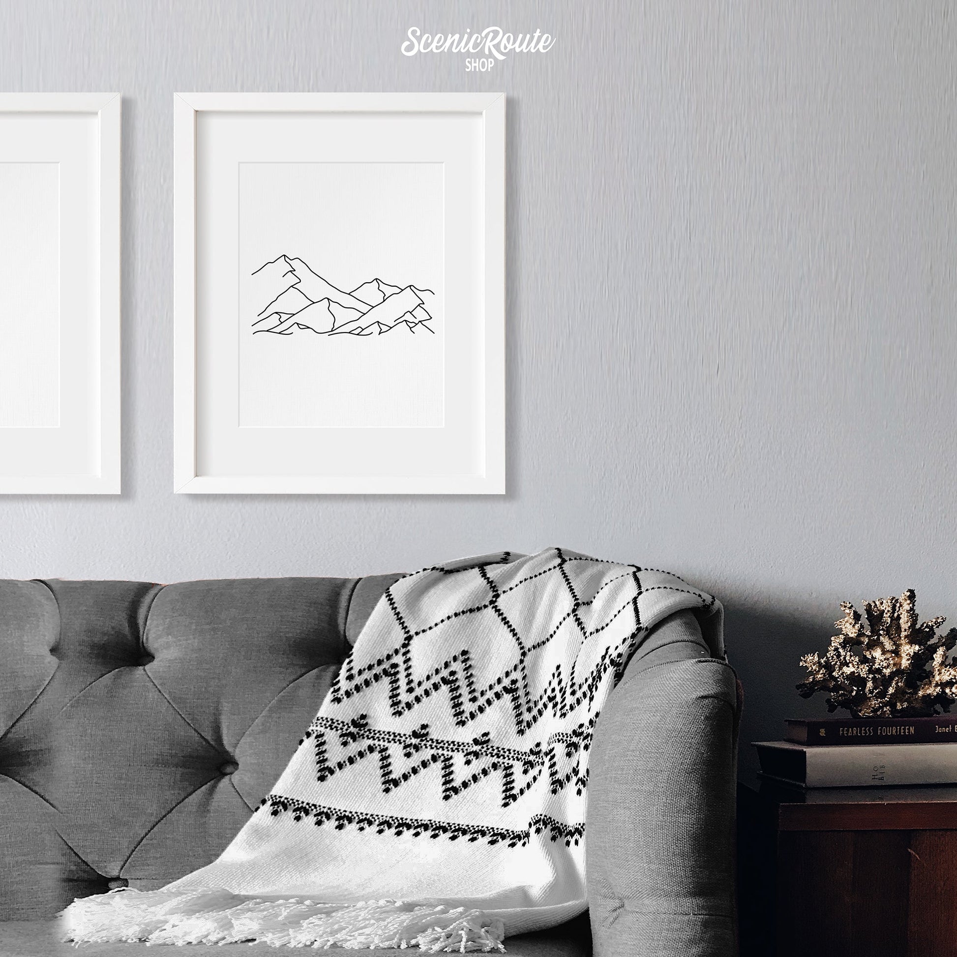 A framed line art drawing of A Mountain Range on a white wall above a couch with a blanket