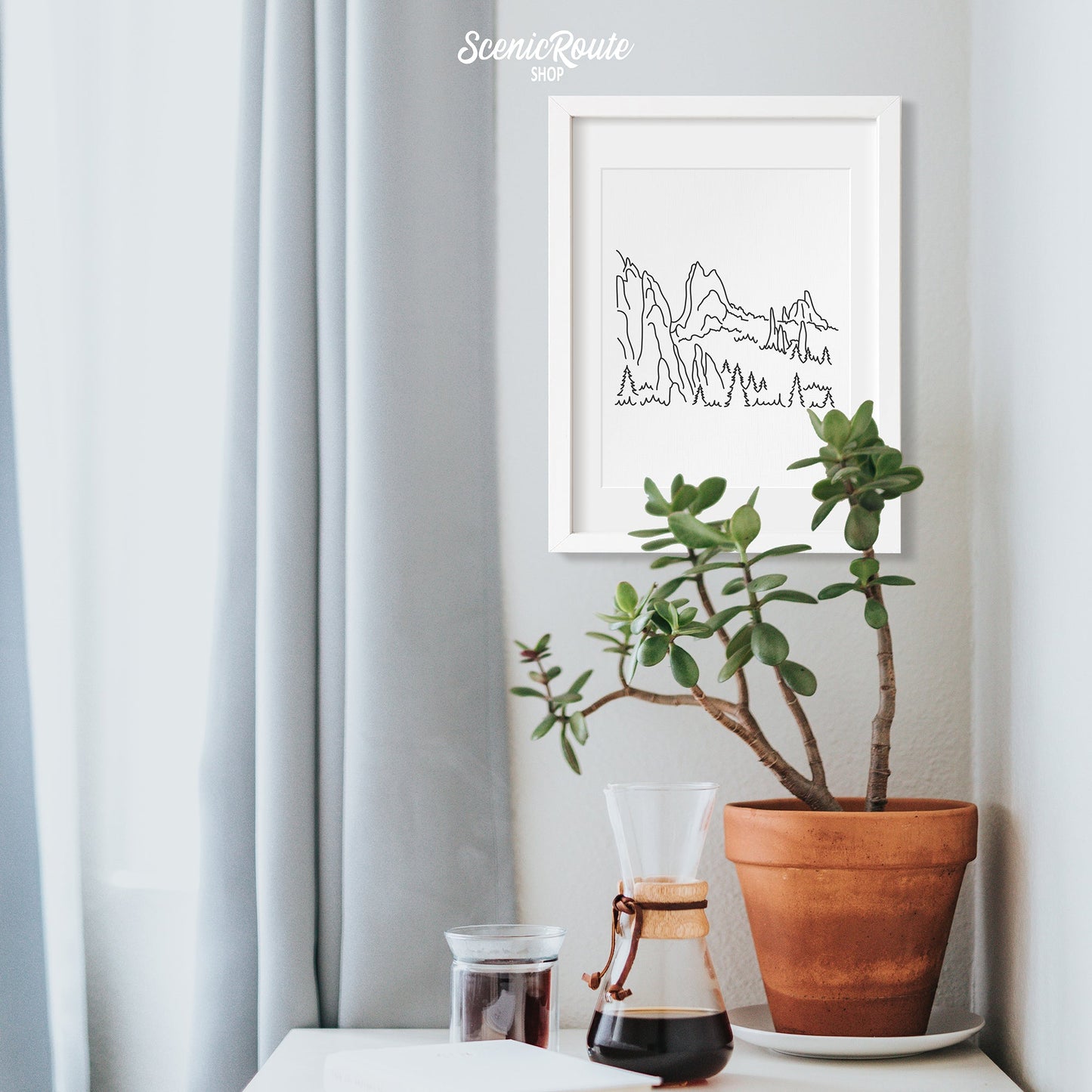A framed line art drawing of The Garden of the Gods hung in a corner above a table with a potted plant